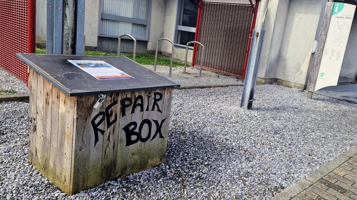 La RepairBox : une caisse en bois cadenassée portant l'inscription « REPAIR BOX »
                  peinte à la main, installée en extérieur près d'arceaux à vélos et d'un panneau « Centre-Ville »